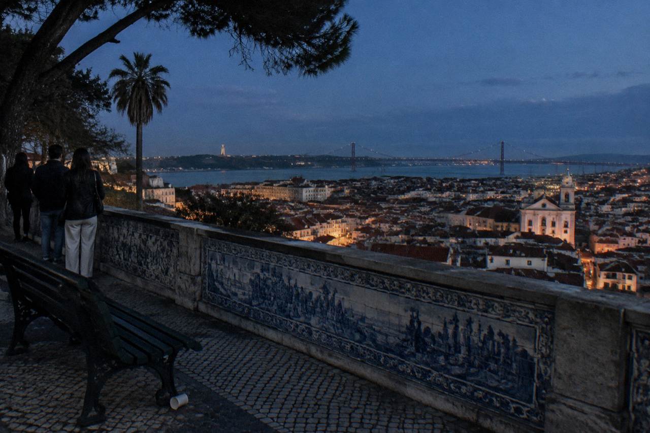 People stand by a tiled terrace overlooking Lisbon rooftops and the Tagus at dusk, with a bridge and lit church visible.