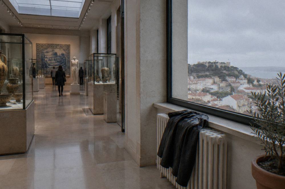 A museum gallery with glass display cases, polished stone floors, visitors, and a large window overlooking Lisbon and the river.