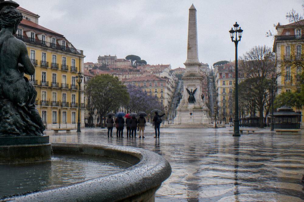 A rainy Lisbon square with a group holding umbrellas near an obelisk and a fountain, surrounded by yellow historic buildings.