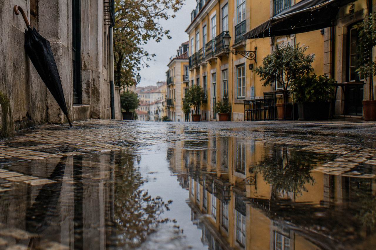 Wet cobblestone street with large puddle reflections, yellow facades, balconies, potted trees, and a black umbrella leaning on a wall.