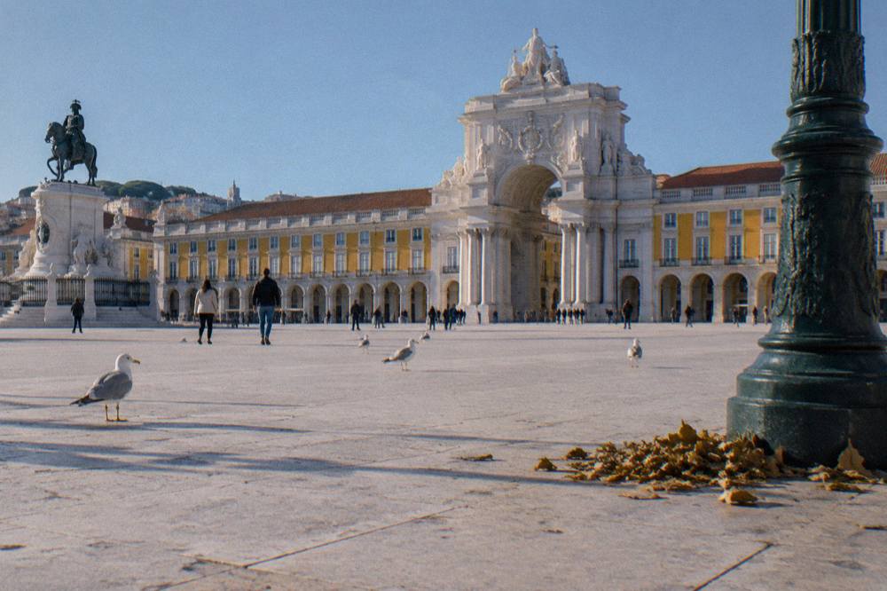 Praça do Comércio with the Rua Augusta Arch, an equestrian statue, gulls on pale paving, and a lamppost in the foreground.