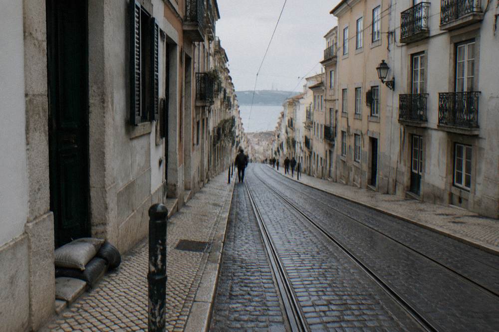 Steep Lisbon street with tram tracks, tall old facades, a few distant pedestrians, and a view down toward the river.
