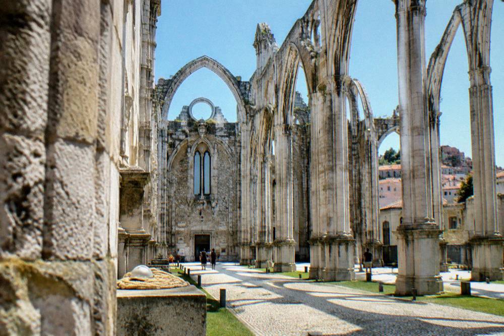 Roofless Gothic convent ruins with tall stone arches, cobbled ground, a hard hat on a ledge, and blue sky overhead.