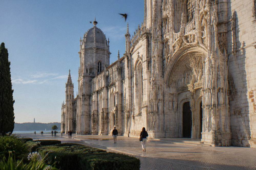 Ornate limestone facade of Jerónimos Monastery beside a waterfront promenade with a few people and distant trees under a clear sky.