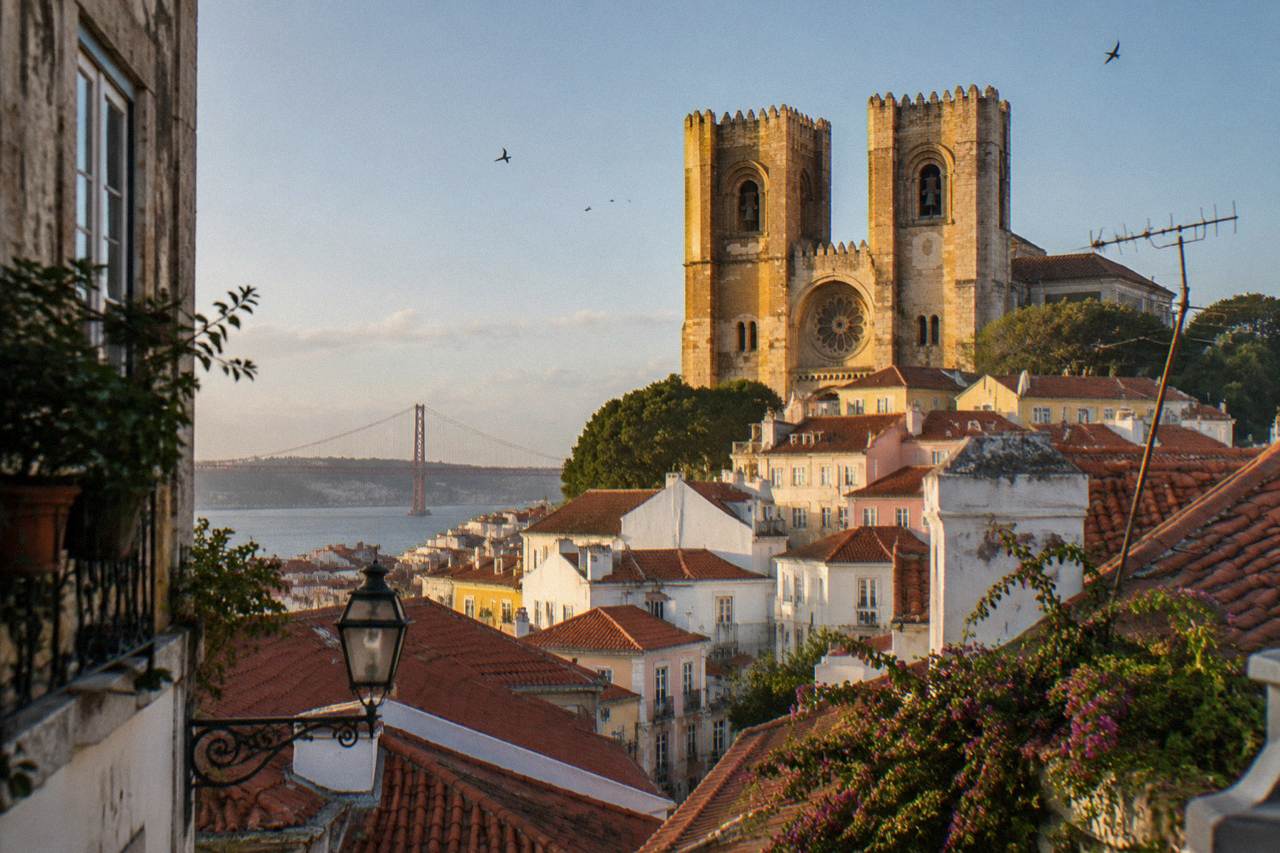 Lisbon Cathedral rises above red-tiled rooftops with the Tagus and a bridge in the distance under warm evening light.