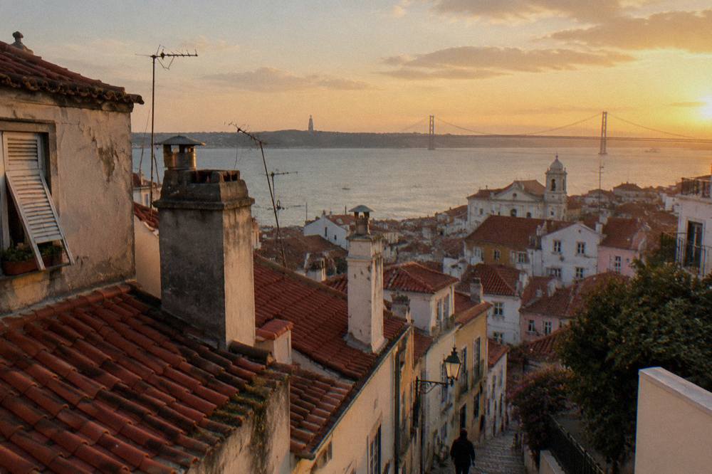 Terracotta rooftops and chimneys step down toward the Tagus at sunset with the 25 de Abril Bridge in the distance.