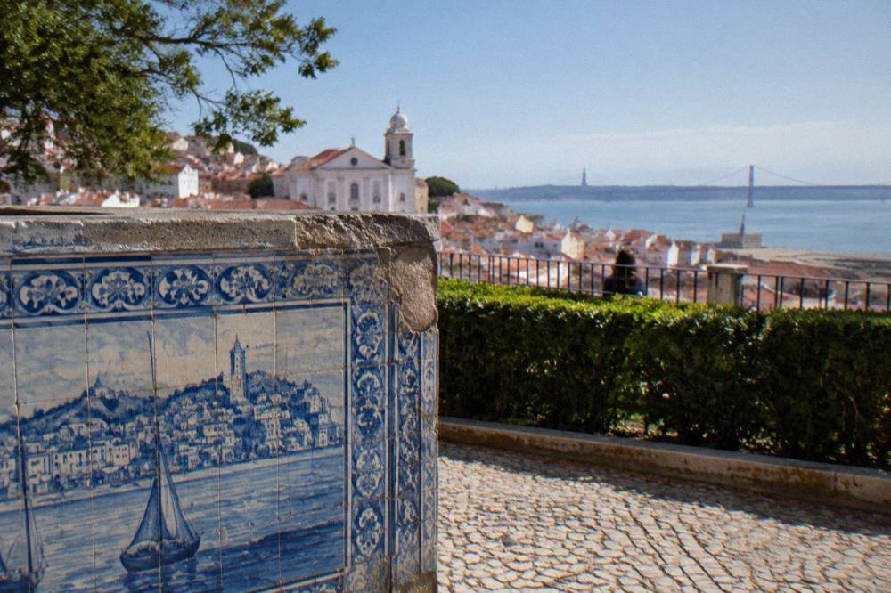 A blue azulejo wall, hedge, railing, cobbled terrace, and Alfama rooftops overlook the Tagus.