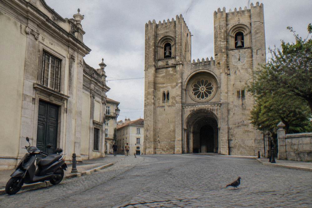 Lisbon Cathedral with twin stone towers fronts a cobbled square, with a parked scooter at left and a pigeon in the foreground.