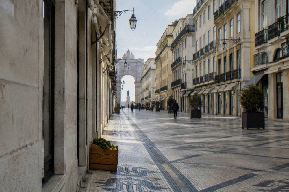 A straight cobbled Lisbon street lined with tall pastel buildings, planters, and a distant triumphal arch over the road.