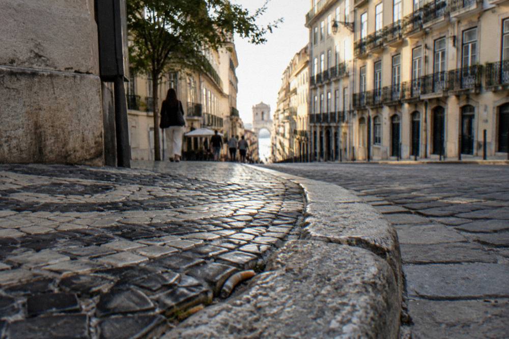 Low-angle view of a Lisbon street corner with black-and-white cobblestones, worn curb stones, and tall pale facades leading downhill.