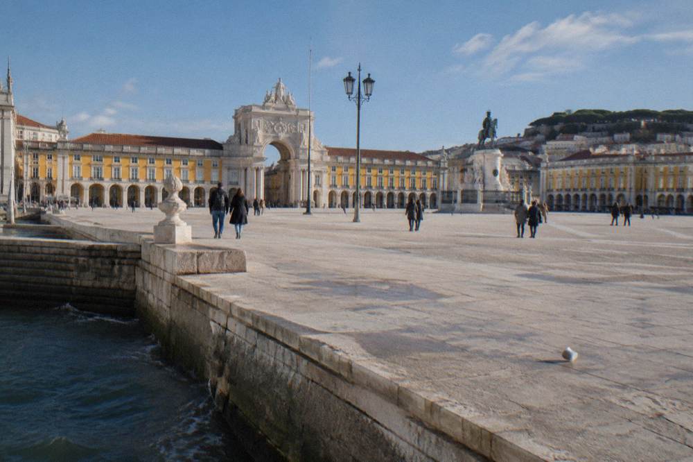 A wide riverside square with yellow arcaded buildings, an arch, an equestrian statue, a few pedestrians, and water at the edge.