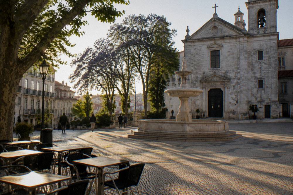 A stone church stands beside a fountain and café tables in a cobbled Lisbon square with tall trees and long shadows.