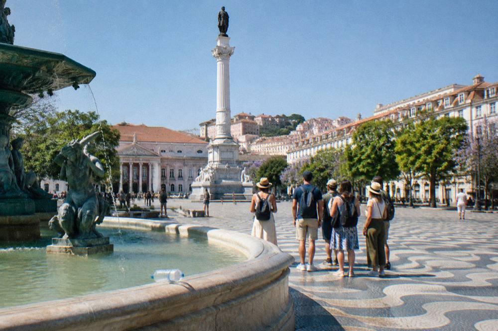 A group of people stands near a fountain and tall column in Rossio Square on a sunny day.