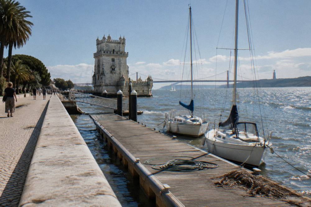 A riverside promenade with moored sailboats, a wooden dock, the Tower of Belém, and the Tagus estuary under a clear sky.