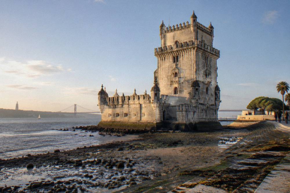 Belém Tower stands beside the Tagus at low tide with mud flats, a riverside path, and the 25 de Abril Bridge in the background.