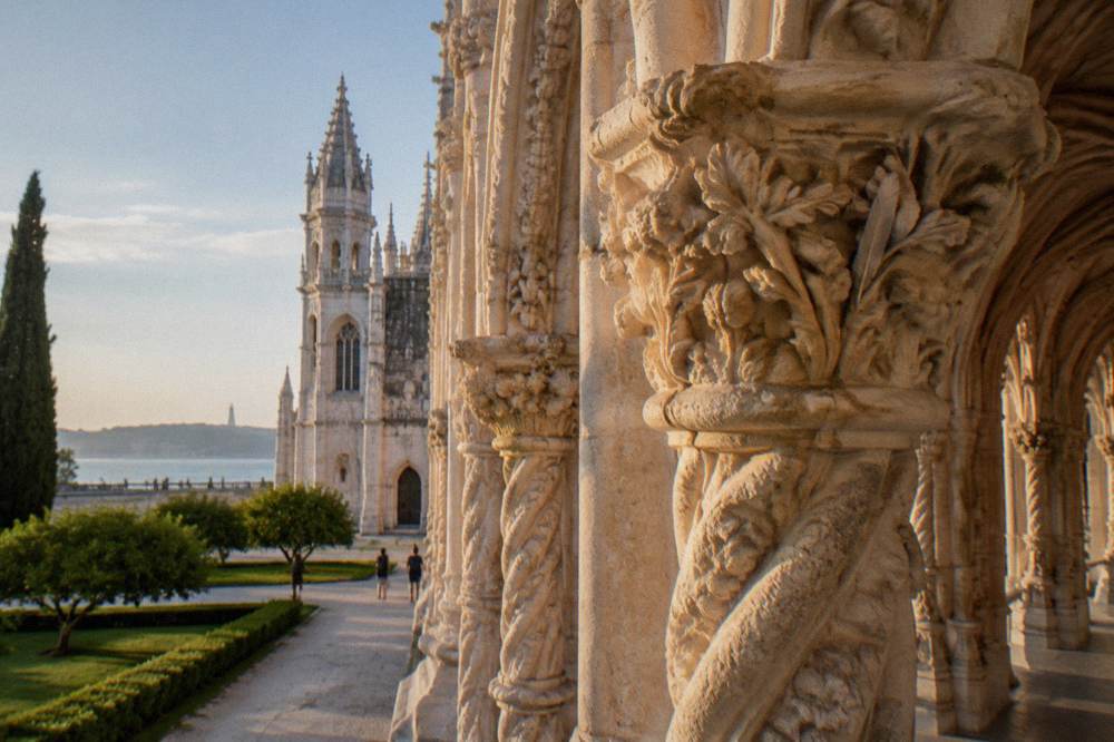 Carved limestone monastery arcade with twisted columns beside the Jerónimos church tower and riverside garden at sunset.