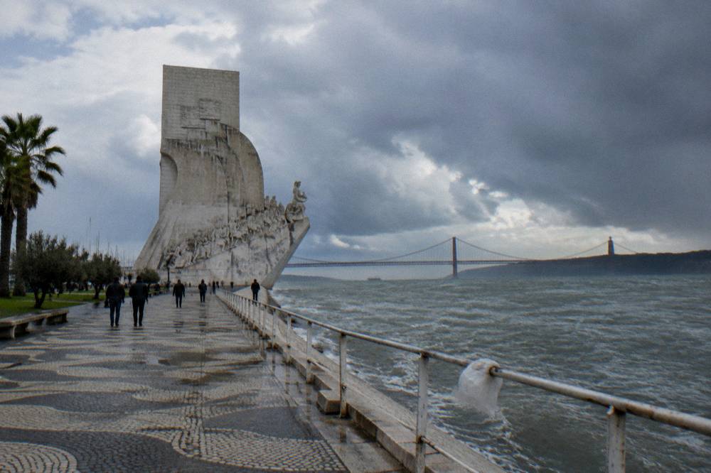 The Monument to the Discoveries stands beside a wet riverside promenade with the Tagus and a suspension bridge under stormy clouds.