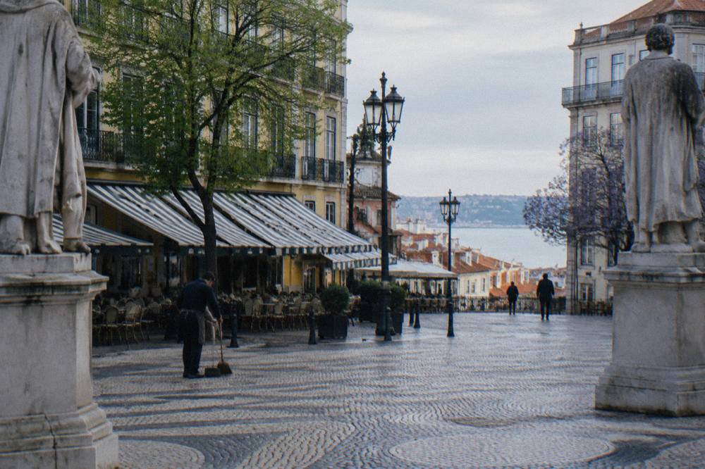 A cobbled Lisbon square with striped cafe awnings, statues, lampposts, and a view down to the Tagus.