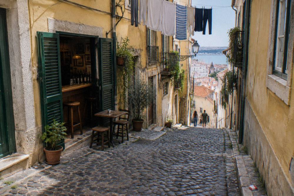 A steep cobbled Lisbon lane with yellow facades, an open bar doorway, laundry hanging overhead, and a distant river view.