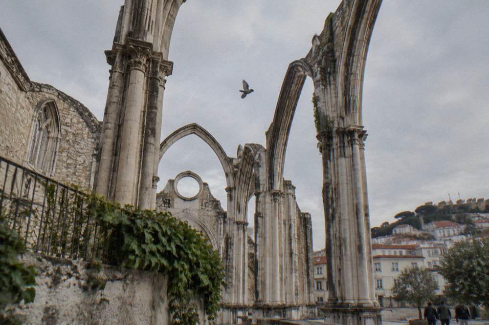 Roofless Gothic arches of a ruined convent rise over a courtyard with a pigeon in the cloudy sky.