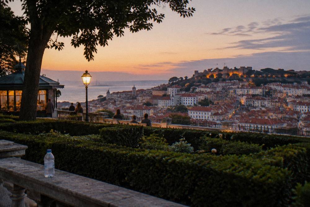 A terraced garden overlook with a lit lamp, hedges, a water bottle on stone balustrade, and Lisbon rooftops and castle hill at dusk.