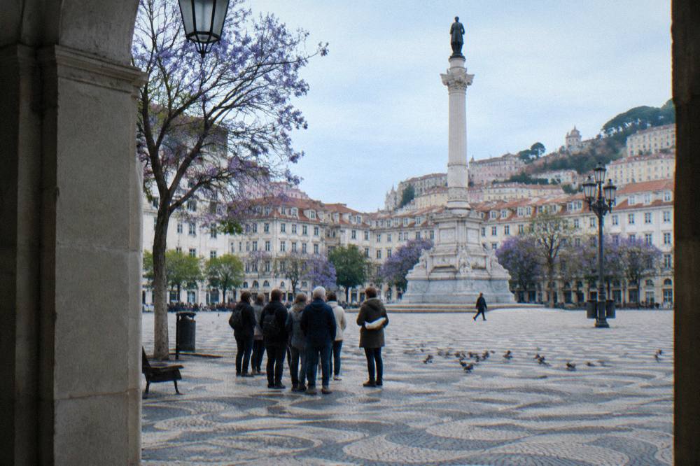 People stand on patterned calçada in a Lisbon square beside a tall monument and purple jacaranda trees under a cloudy sky.