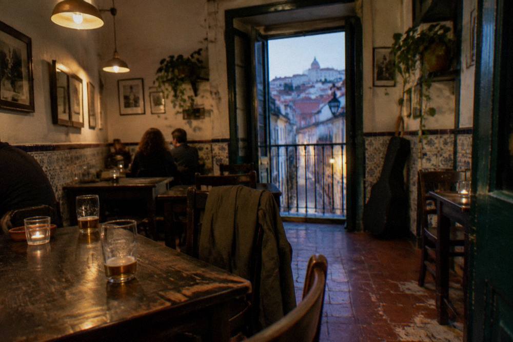 A dim tasca interior with wooden tables, hanging lamps, tiled walls, beer glasses, seated patrons, and a guitar case by a doorway.