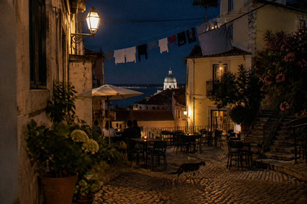 A lit cobbled square with café tables, washing lines, a cat crossing, and rooftops overlooking the river at night.