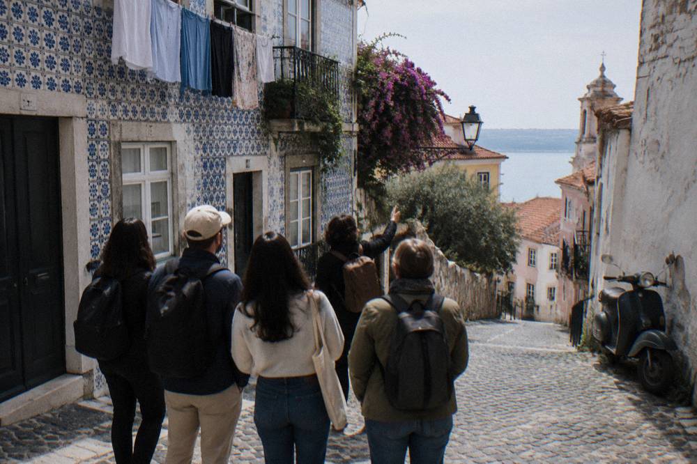 Five people walk down a cobbled Alfama street beside blue-tiled facades, laundry, flowering vines, and a parked scooter.