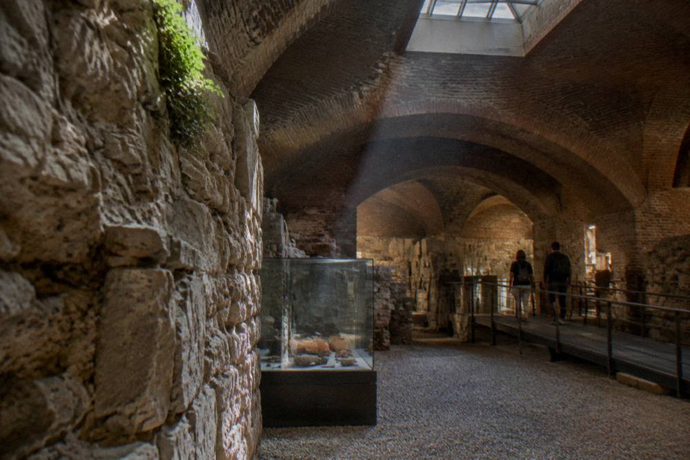 An underground brick-arched gallery with stone walls, a skylight, display cases, and two visitors on a walkway.