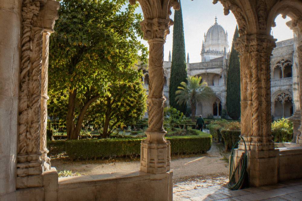 Ornate stone cloister arcade frames a sunlit garden with trimmed hedges, tall cypress trees, and a distant person.