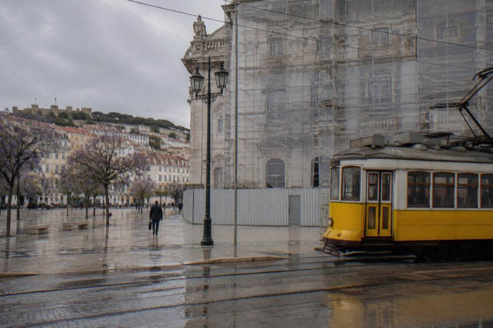 A yellow tram stands beside a scaffolded pale stone building in a wet Lisbon square with purple jacaranda trees and hillside buildings.