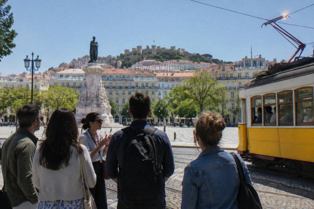 A guide speaks to a small group in a Lisbon square beside tram tracks, with a yellow tram and castle hill in the background.