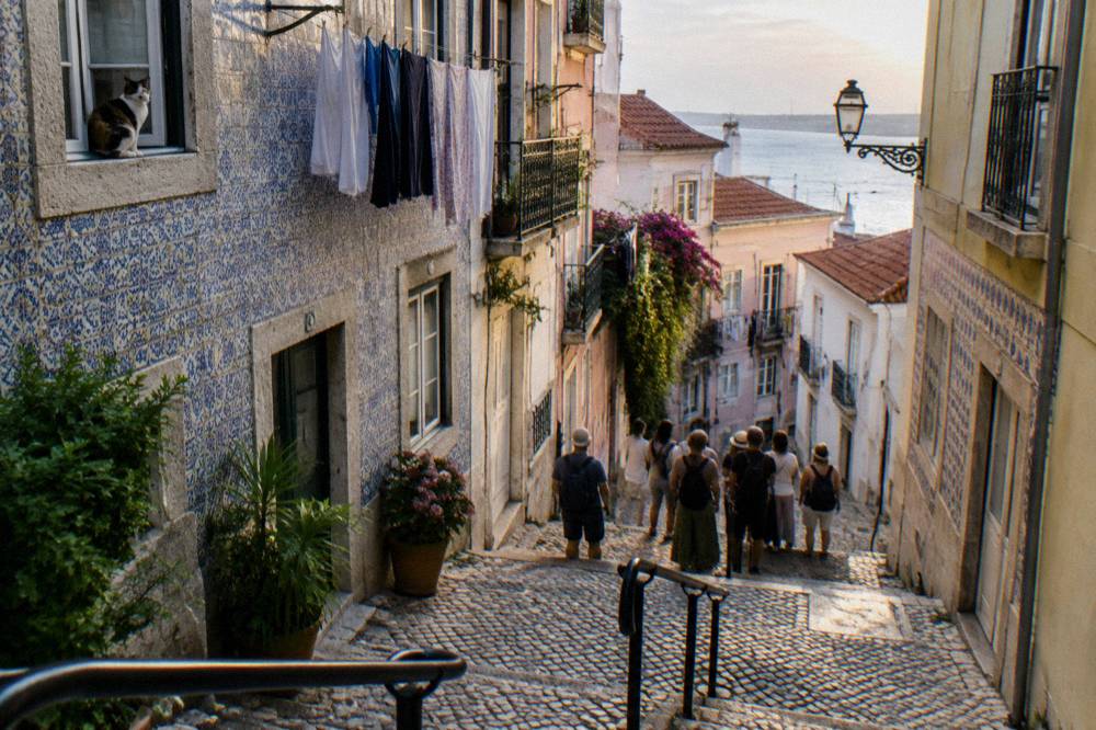A small group stands on a steep cobbled street between azulejo-covered apartment buildings, with a cat in a window and the river beyond.