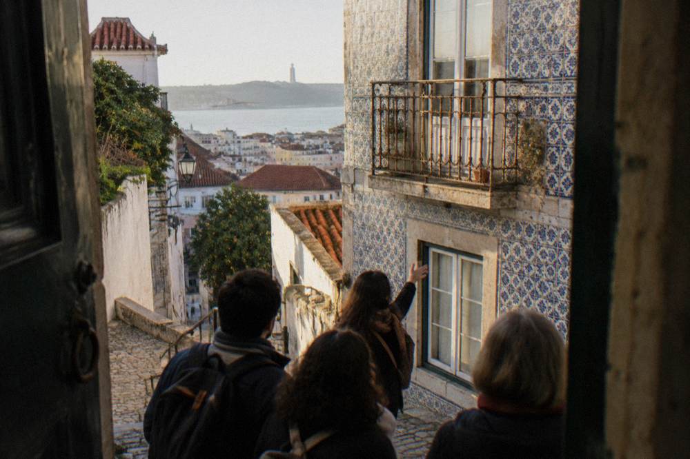 Several people stand on a narrow cobbled Lisbon lane beside a blue-tiled facade, with a guide gesturing toward the wall and the Tagus visible beyond.