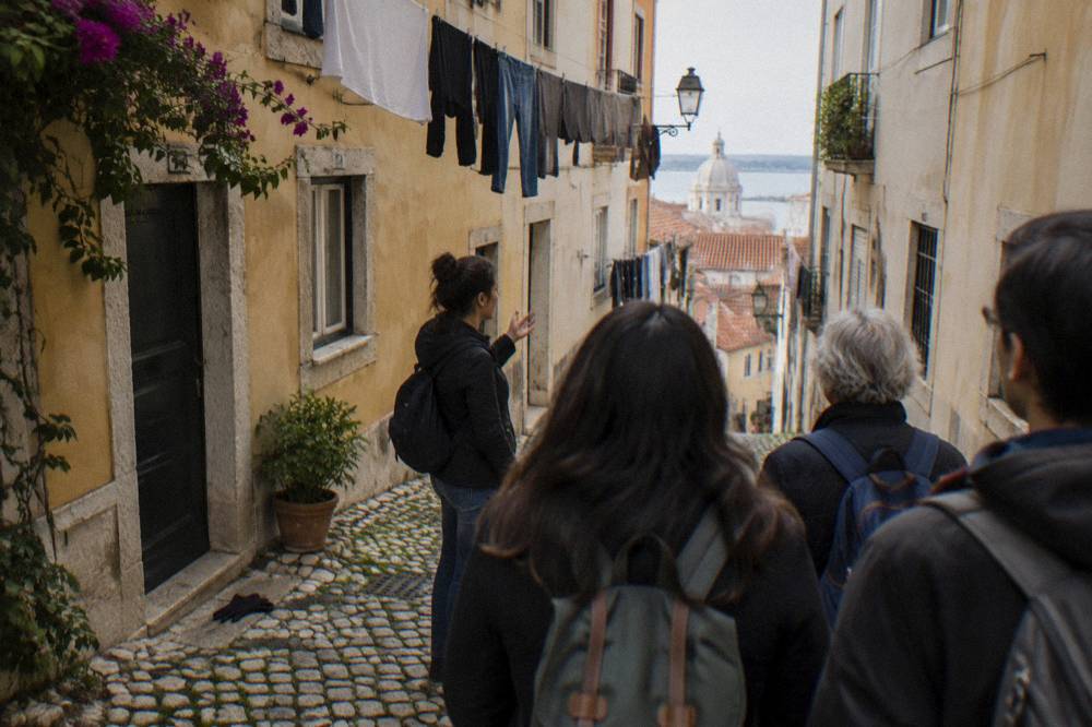 A group of walkers follows a guide down a narrow cobbled Lisbon street with laundry hanging overhead and the Tagus visible beyond.