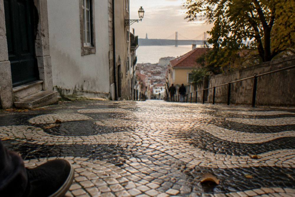 Wet black and white patterned cobblestone street slopes downhill between buildings toward the river and bridge.