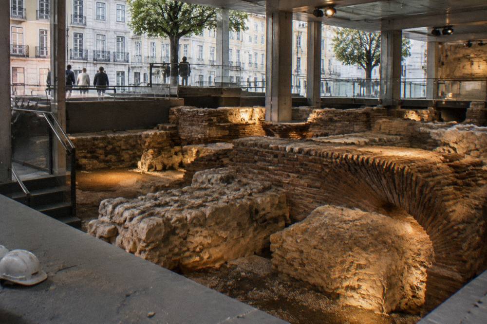 Underground archaeological ruins with stone and brick foundations, warm spotlights, a hard hat on a ledge, and street buildings visible above.