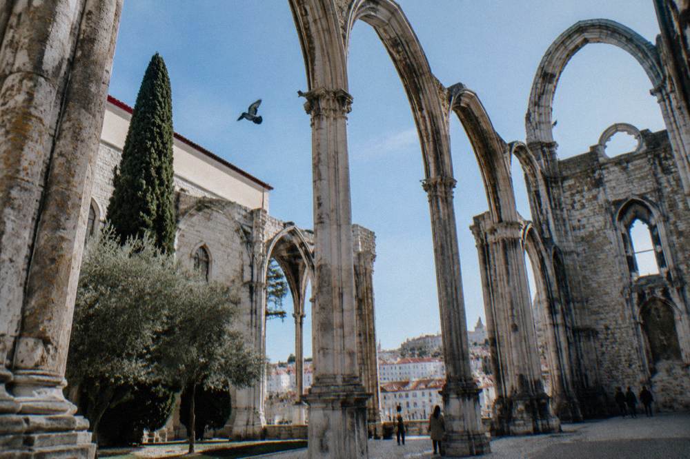 Roofless Gothic stone arches of ruined Carmo Convent rise over a Lisbon courtyard with a pigeon in the sky and hillside buildings beyond.
