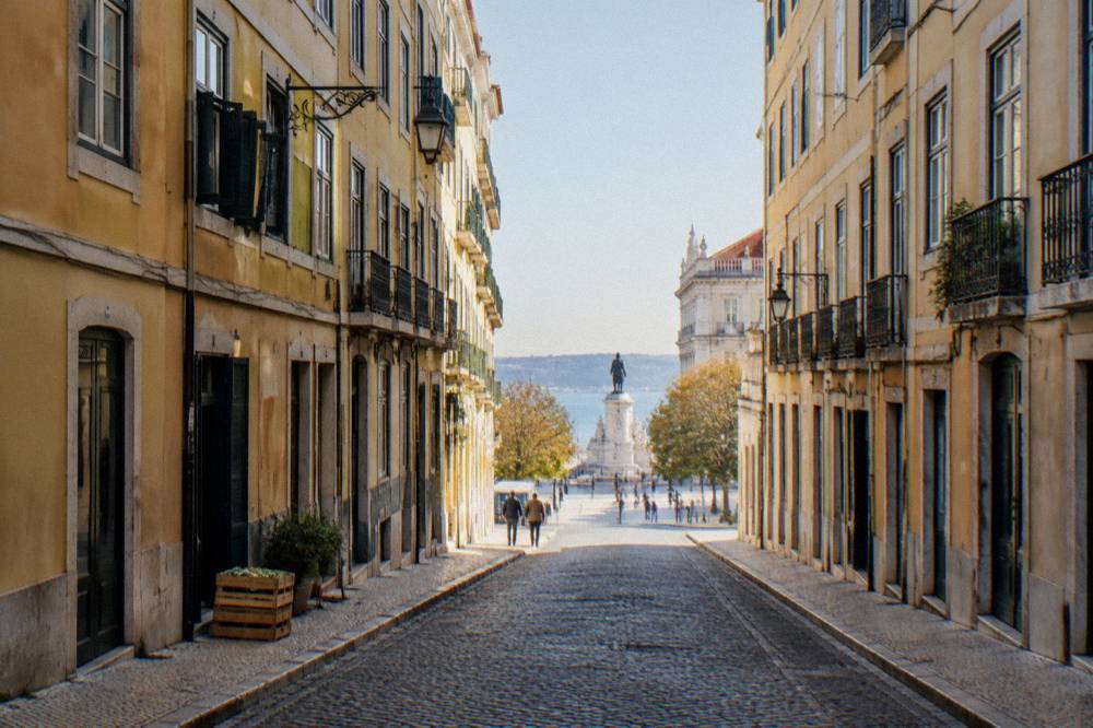 A cobbled Lisbon street lined with yellow facades leads down to a sunlit square and a riverside statue.