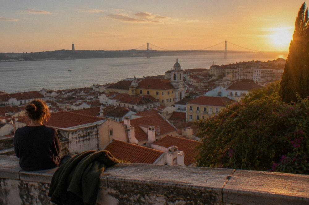 A person sits on a stone parapet above terracotta rooftops with the Tagus and 25 de Abril Bridge at sunset.