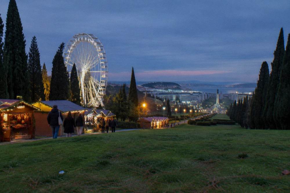 Festive market stalls and a lit ferris wheel line a grassy Lisbon park avenue at dusk with cypress trees and city views.