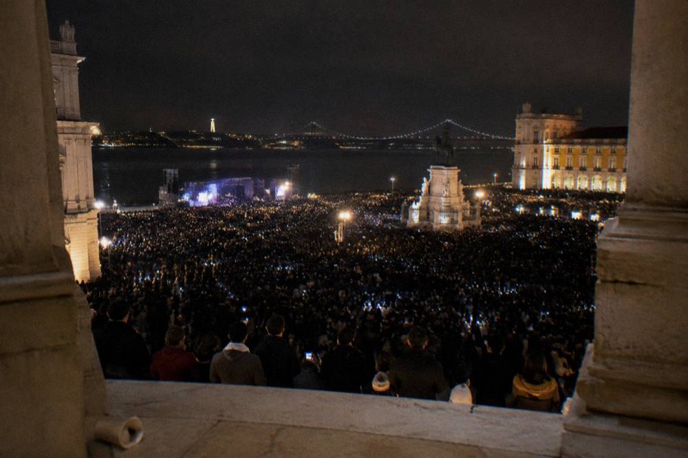 A huge crowd fills Praça do Comércio at night beside the river, viewed from a stone arcade with glowing phones and distant lights.