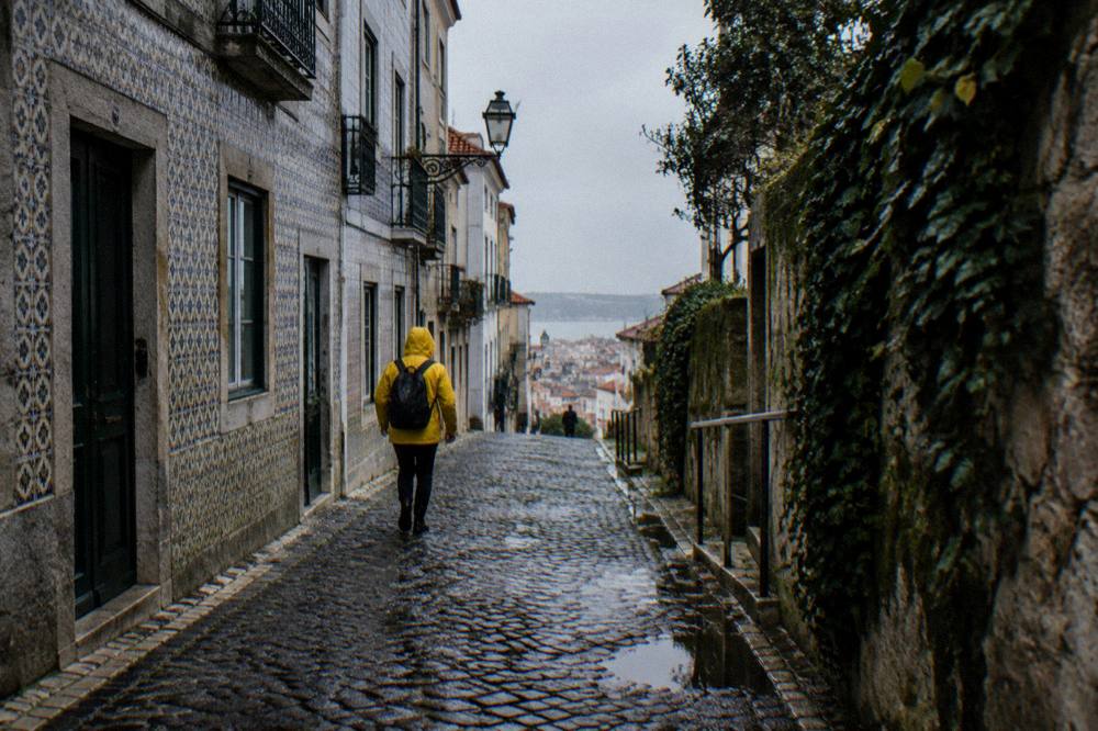 A person in a yellow rain jacket walks down a wet cobbled Lisbon street between tiled buildings under a grey sky.