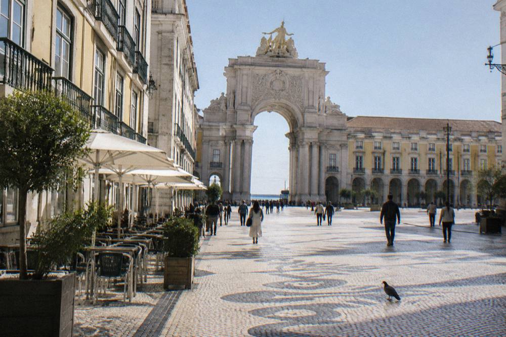 A wide cobbled Lisbon plaza leads toward the Rua Augusta Arch with people walking, café tables on the left, and a pigeon on the ground.