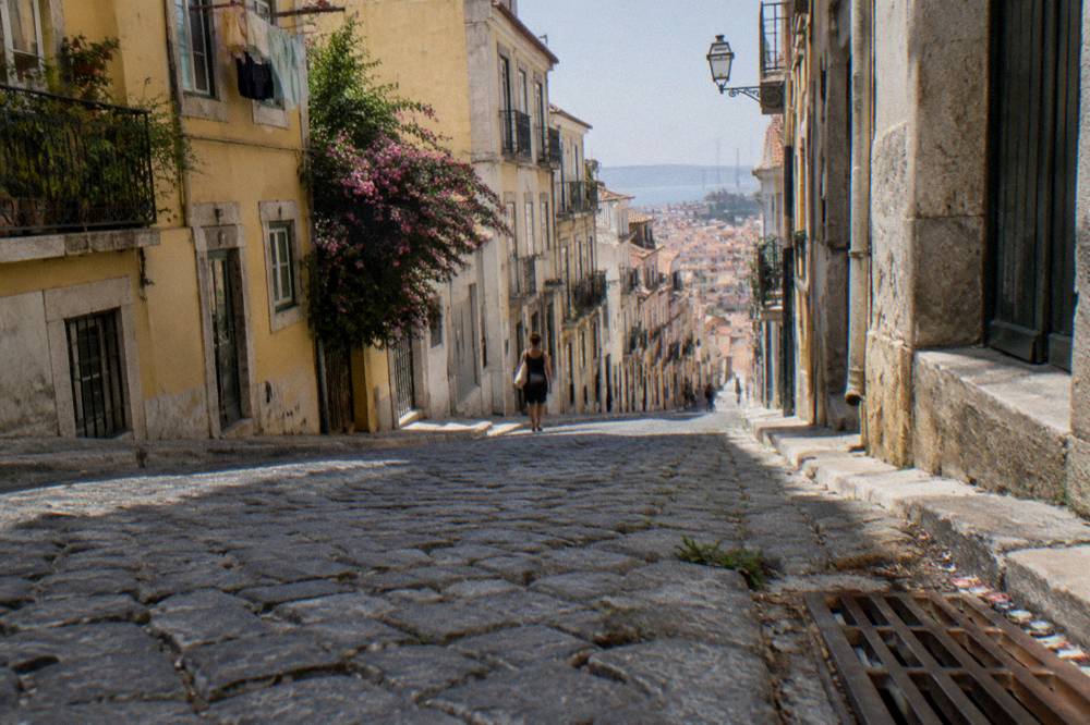 Steep cobbled street lined with old buildings, a flowering balcony, one distant pedestrian, and the river visible downhill.