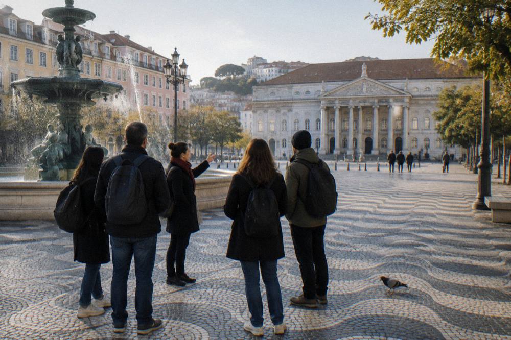 A small group stands on wavy mosaic paving by a fountain in a Lisbon square with a neoclassical theater façade behind them.