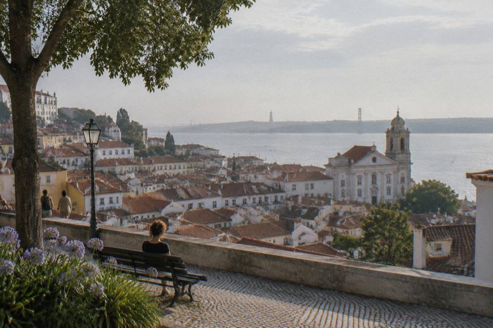 A stone terrace with a bench overlooks tiled rooftops, a white church, and the Tagus under hazy late-afternoon light.