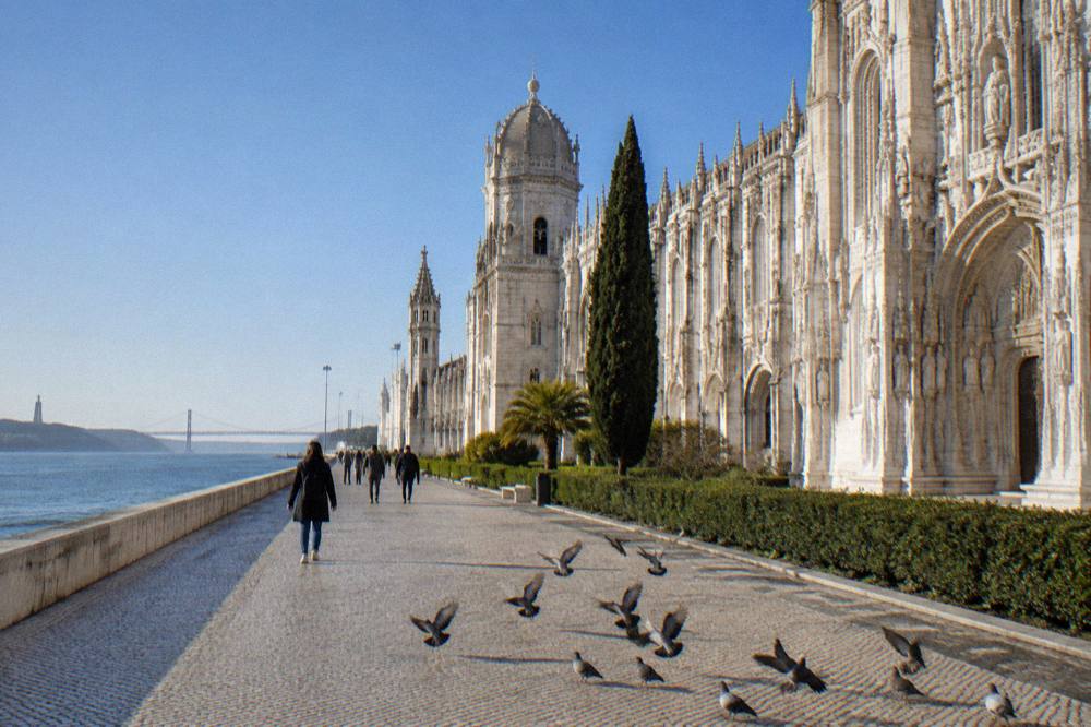 Stone monastery facade lines a riverside promenade with pigeons, walkers, and blue water under a clear sky.