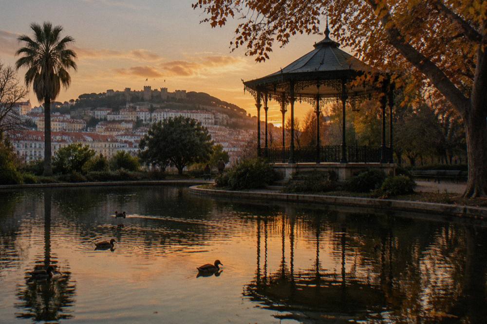 A wrought-iron bandstand stands beside a reflective garden lake with ducks and a sunset view over Lisbon’s hillside homes.